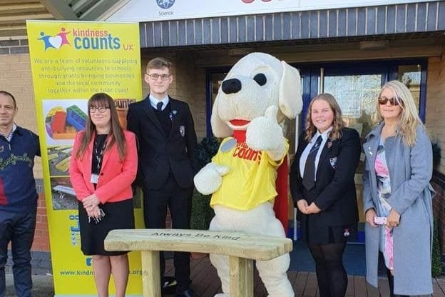 A new buddy bench with “Always Be Kind” carved into it, outside a school, with Nic the mascot and pupils behind it.