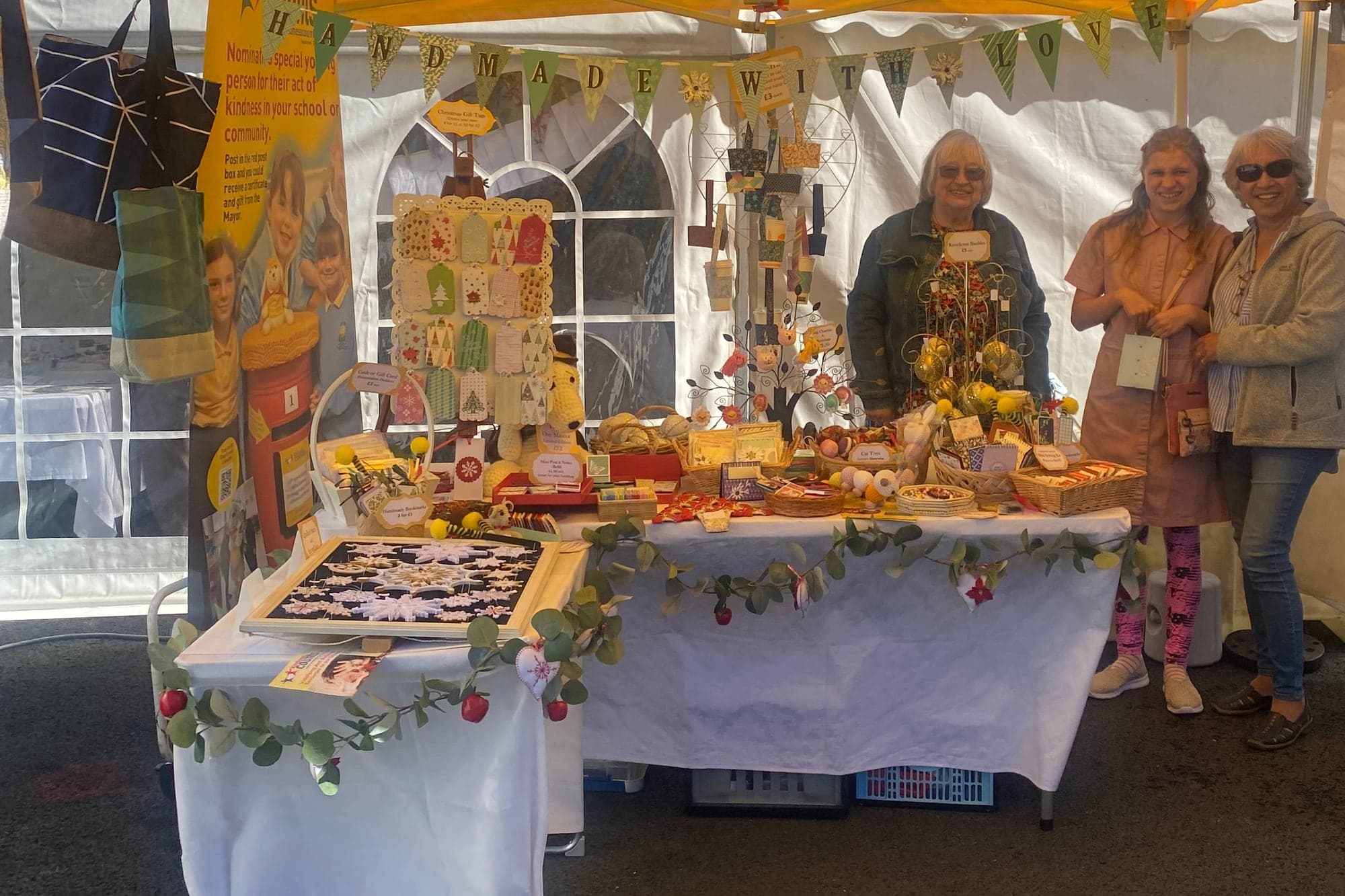 The Kindness Counts UK craft stall at Wrea Green — handmade gifts on a table under a yellow gazebo, with handmade-with-love bunting and three of the crafting team.