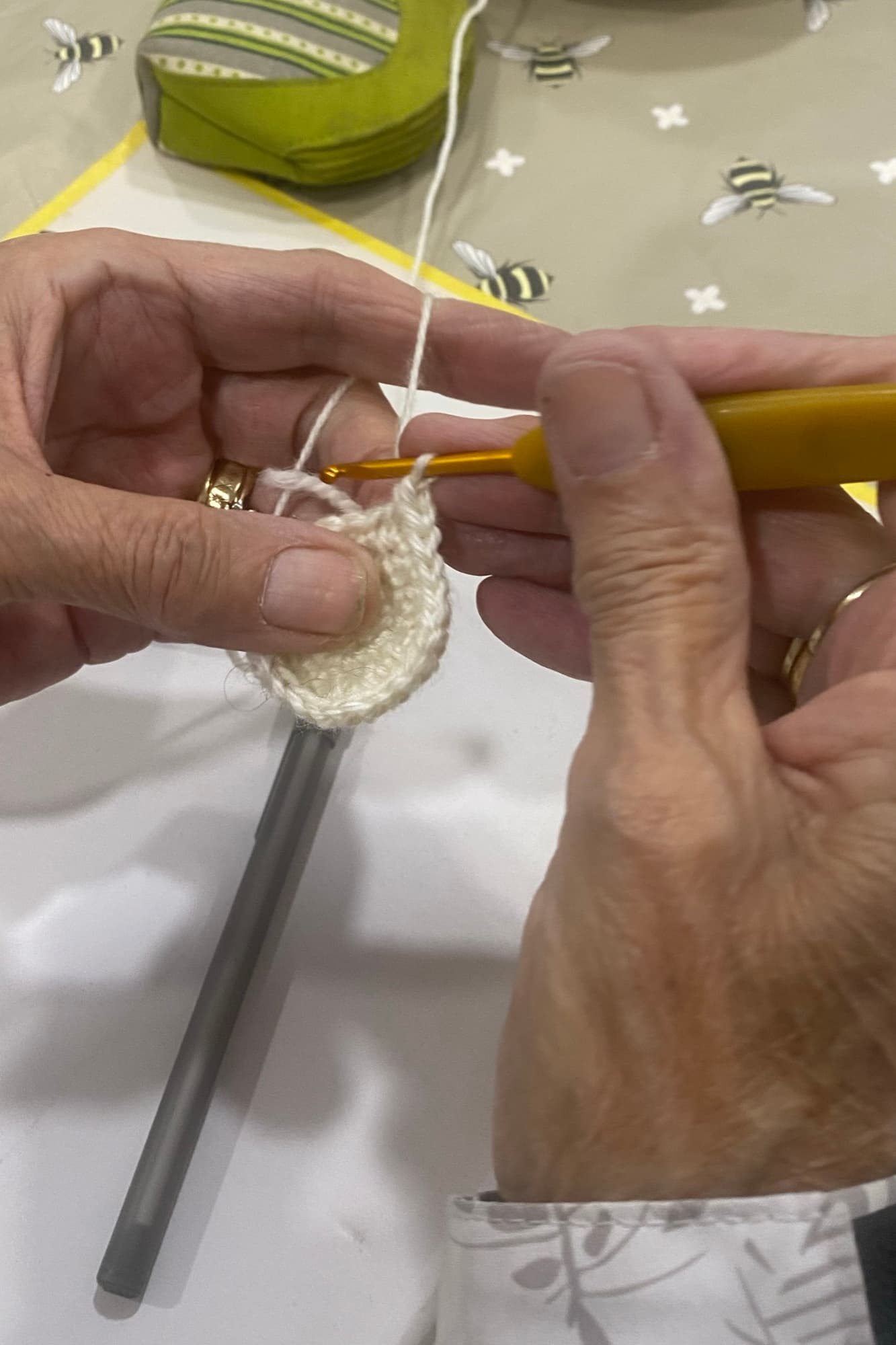 Hands crocheting with a yellow hook at crochet club, a pattern sheet on the table.