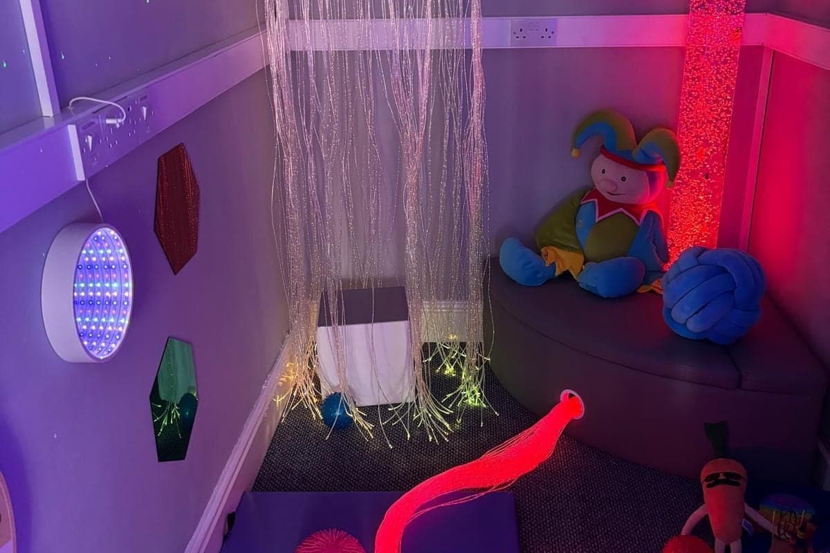 The sensory room, with a fibre optic curtain, a bubble tube and soft seating, lit in purple and red.