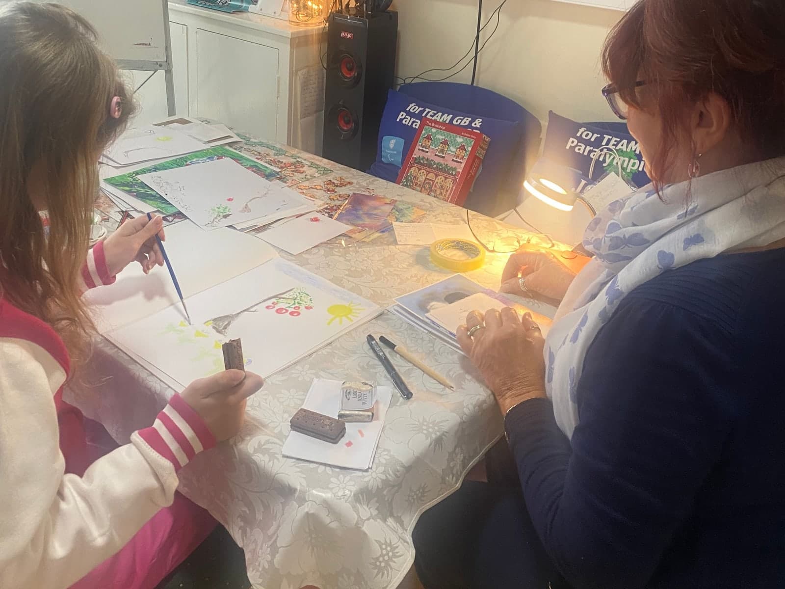 A child painting a tree in watercolours at the Wednesday art class in Hub 1, with Diane of Artydi sitting alongside and artwork spread across the table.