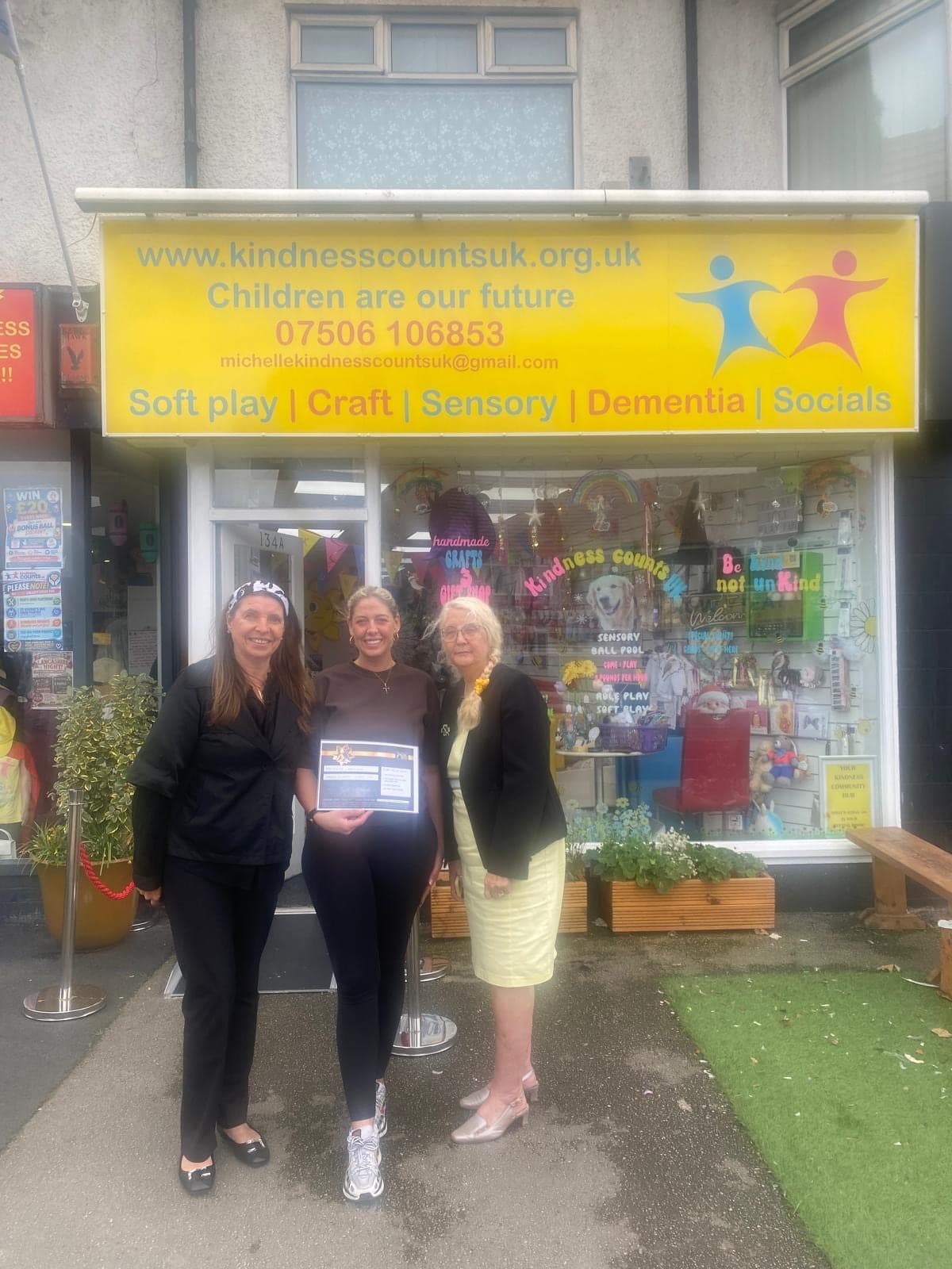 Three people outside the hub on Victoria Road West holding a framed certificate, under the Kindness Counts UK sign.
