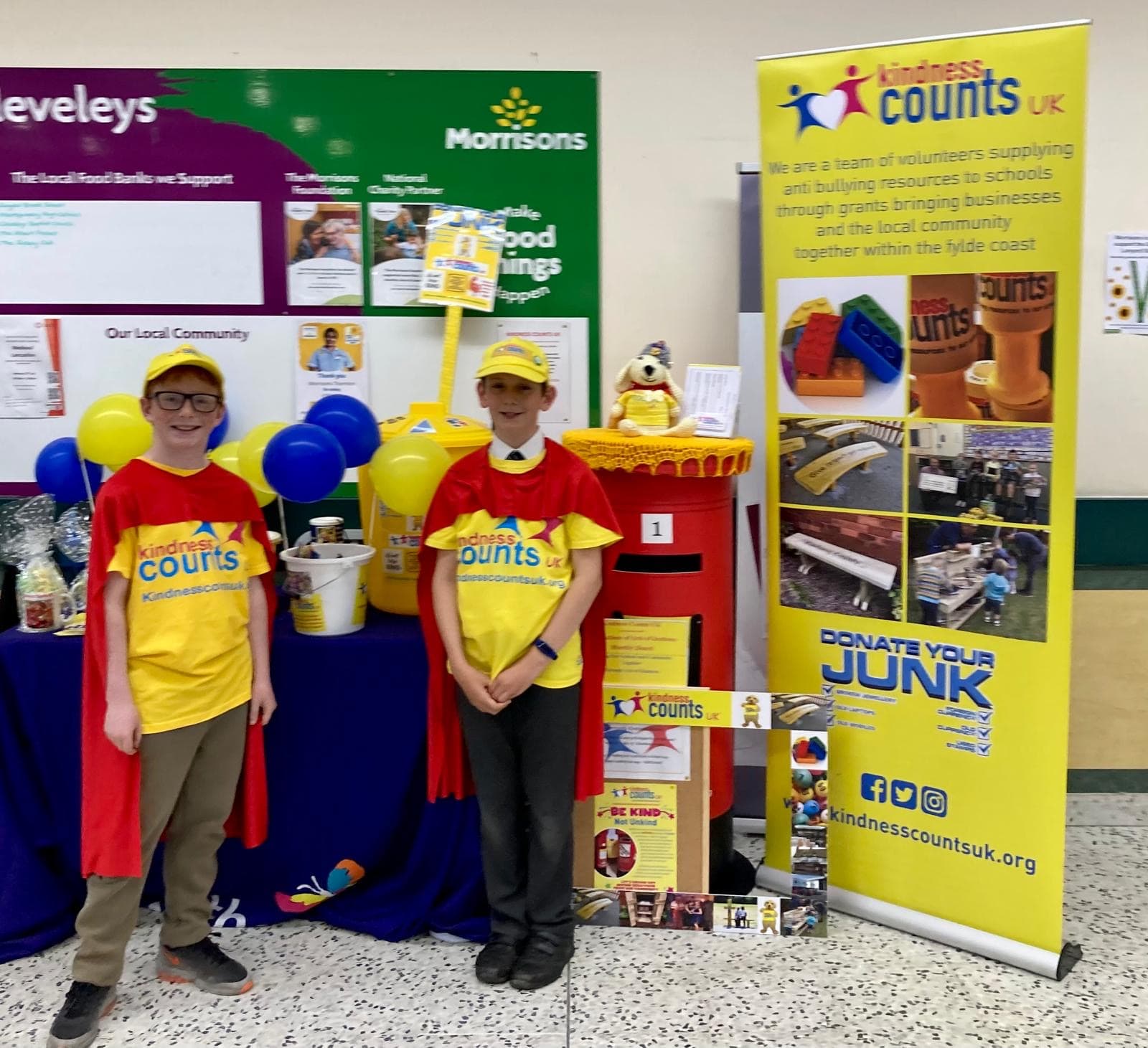 Two young volunteers in Kindness Counts UK t-shirts and red capes beside the kindness post box at a community event in Morrisons, Cleveleys.