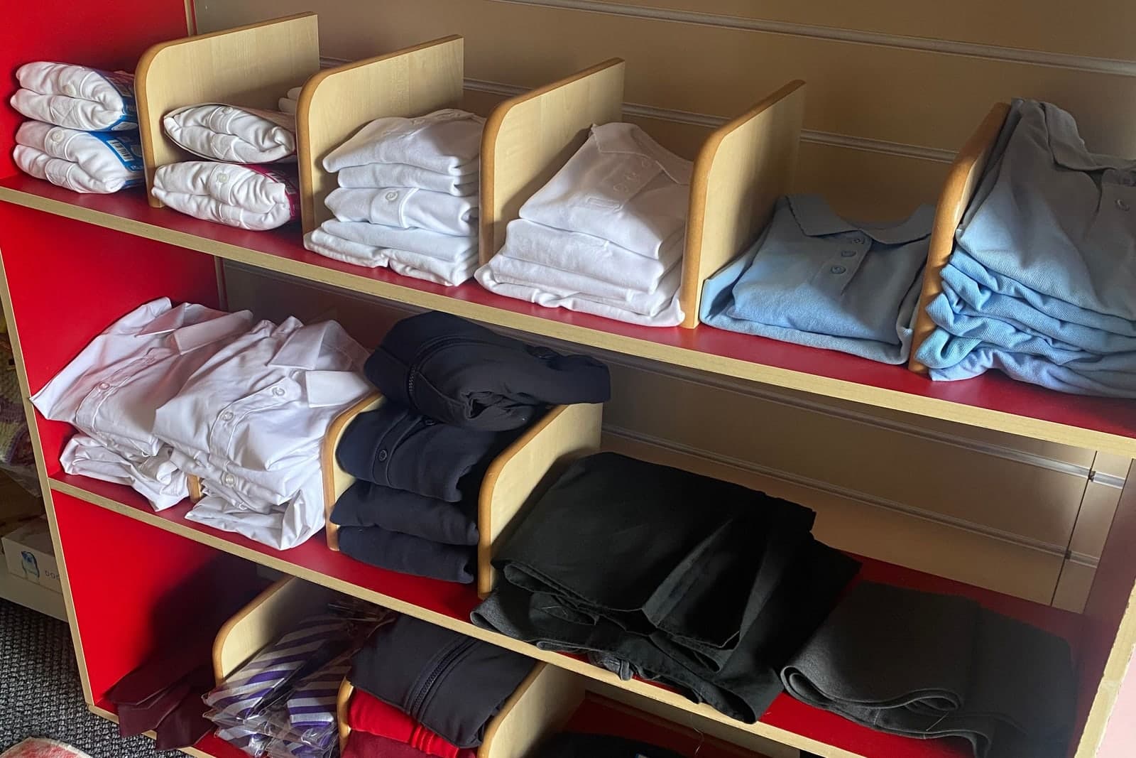 School uniform folded on the shelves at the hub — white and pale blue polo shirts, white blouses, navy jumpers, black trousers and red cardigans.