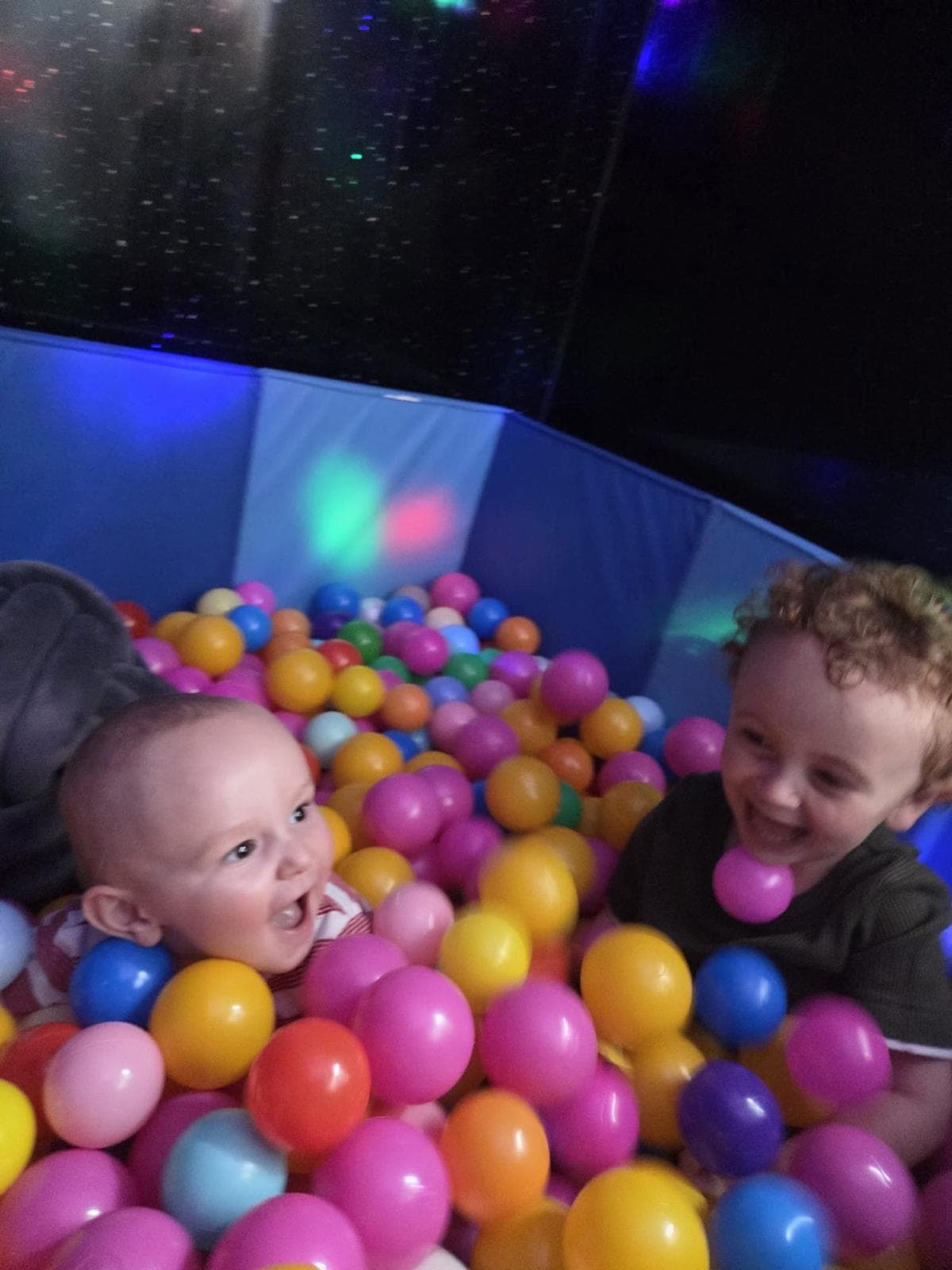 Two small children laughing together in our sensory ball pit, under the coloured lights.