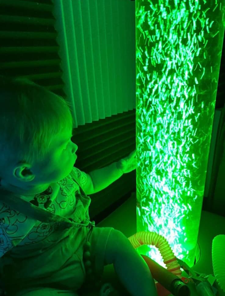 A toddler reaching out to touch the glowing green bubble tube in our sensory room.